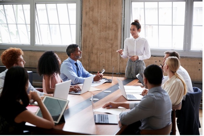 A group of people in professional clothing are sitting around a conference table, listening to a speaker.