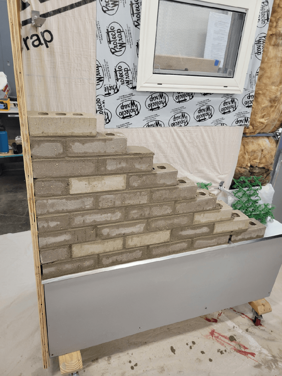 Construction wall mockup with stepped gray brickwork, a screened window, building wrap, exposed insulation, and unfinished framing.