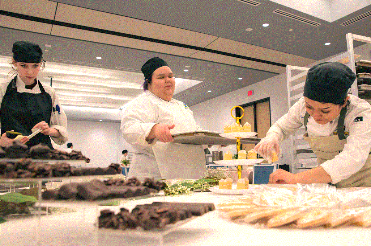 Three student chefs arrange pastries in a state of the art kitchen classroom.