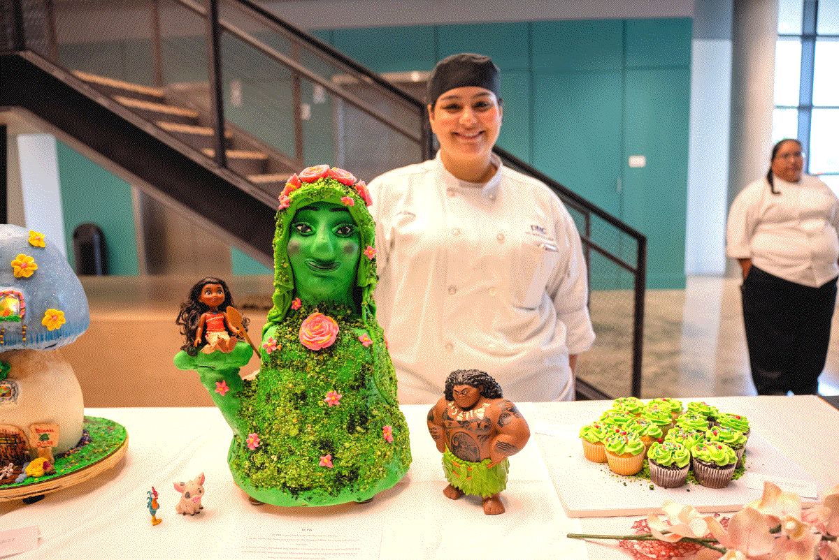 A student chef stands behind a themed cake and cupcakes on a table. The cake is a green character with floral decorations, accompanied by smaller character figures and cupcakes with green frosting.