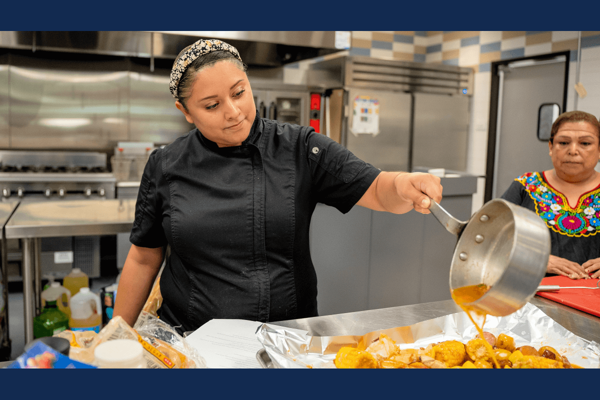 Student in a black chef coat pours sauce over vegetables in a professional kitchen classroom.