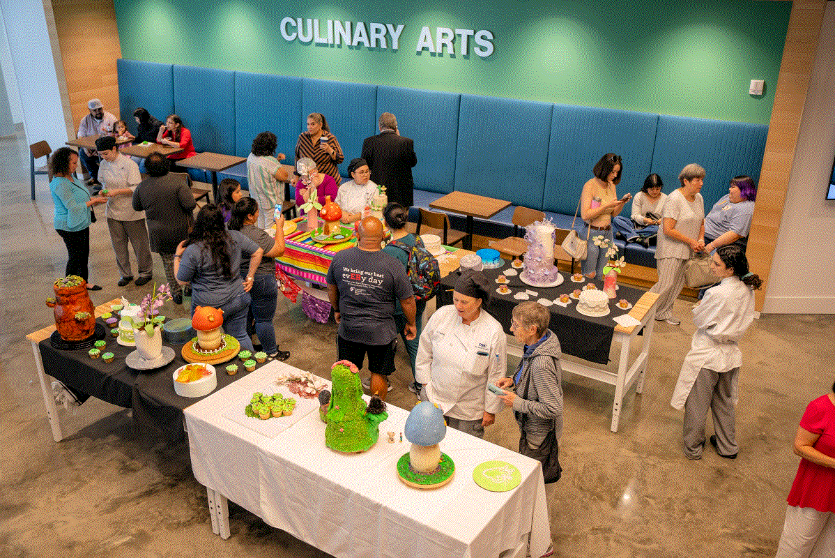 A culinary arts event at the Culinary building with people mingling around tables displaying colorful, decorative cakes and pastries, under a teal wall with "CULINARY ARTS" written on it.