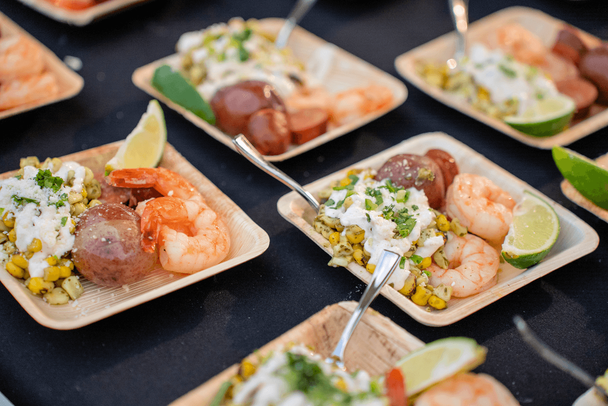 Wooden plates with shrimp, sliced potatoes, corn with herbs, sauce, and lime wedges on a dark tablecloth.