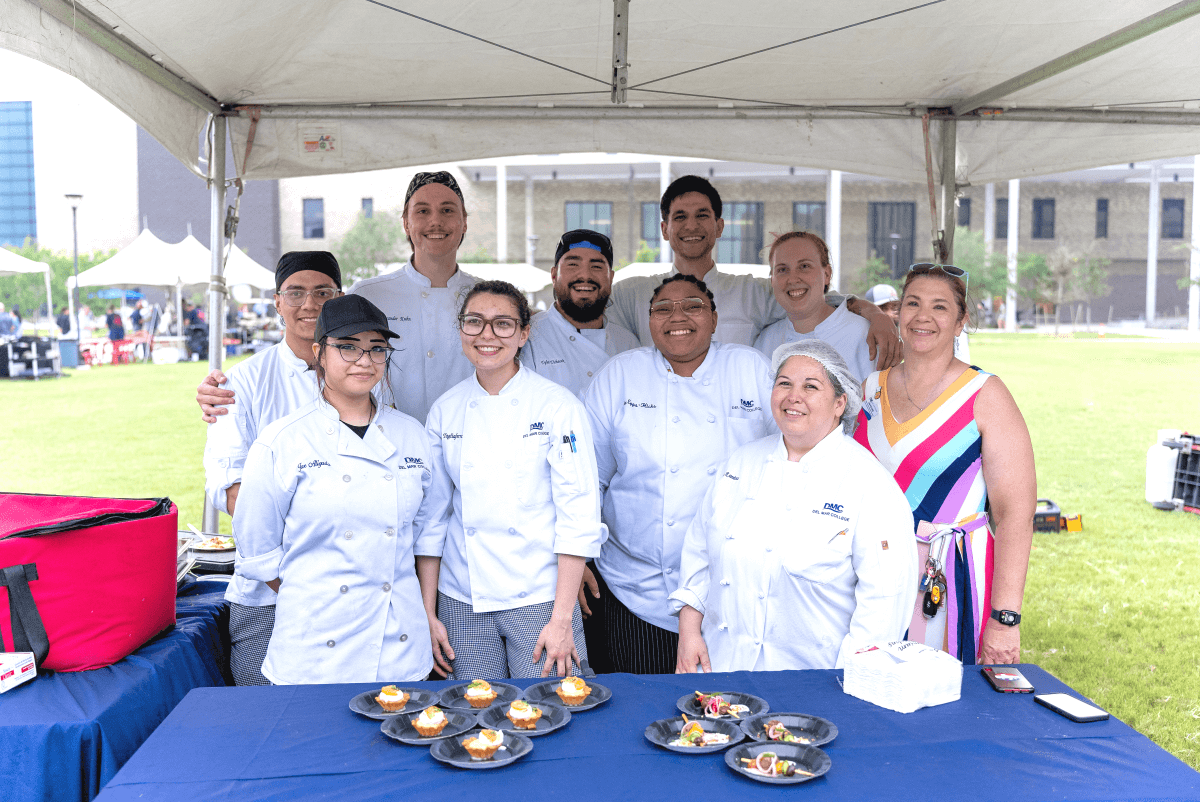 A group of students with Culinary Professor Lisa Pollakis, mostly in chef jackets, standing under a tent with food samples on a table at an event at the Oso Creek Campus.