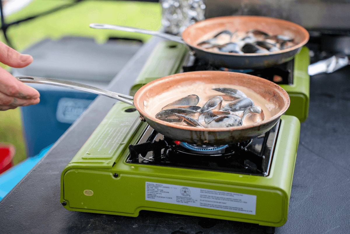 Two frying pans with mussels cooking on portable green gas stoves outdoors at an event at Oso Creek campus.