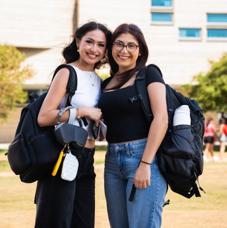 Two young women standing and smiling outdoors, each carrying a black backpack. One wears a white crop top and black pants, while the other is in a black fitted shirt and blue jeans.