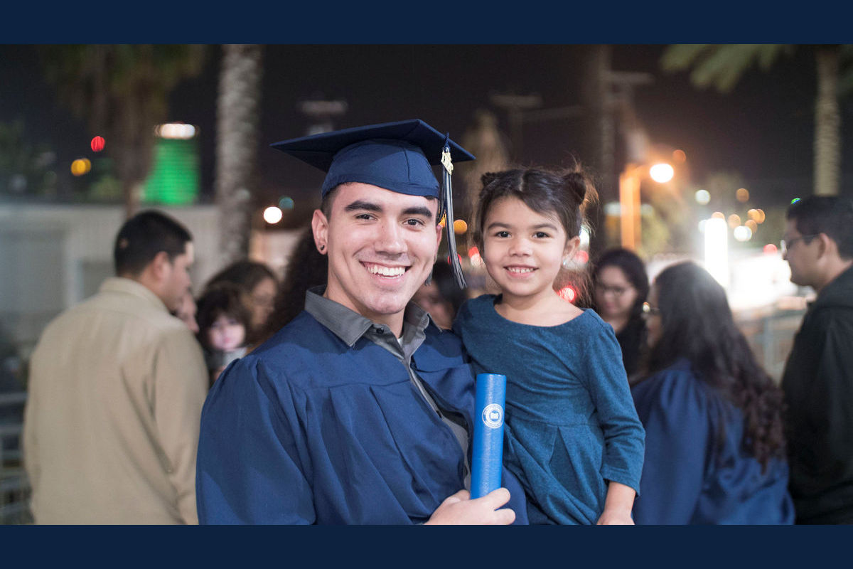 A smiling student poses in cap and gown while holding his degree and a young child
