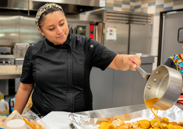 A chef pours sauce over food in a professional kitchen.