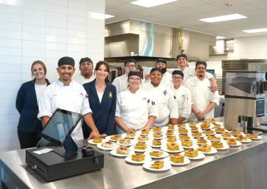 A group of students in a hotel kitchen standing behind a counter lined with plated dishes.
