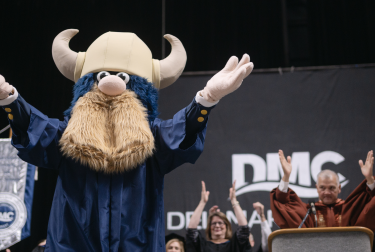 Viking mascot in a horned helmet and blue robe raises both arms onstage as people stand behind it.