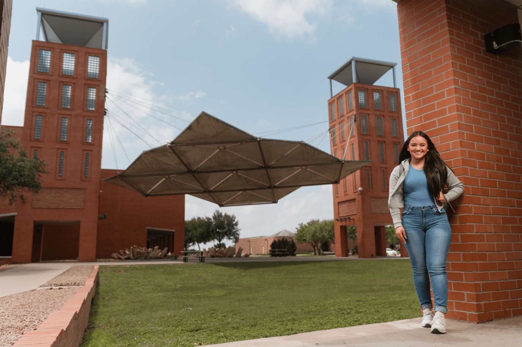 Smiling woman standing beside a brick column in a campus courtyard with red-brick buildings and a large suspended shade canopy.