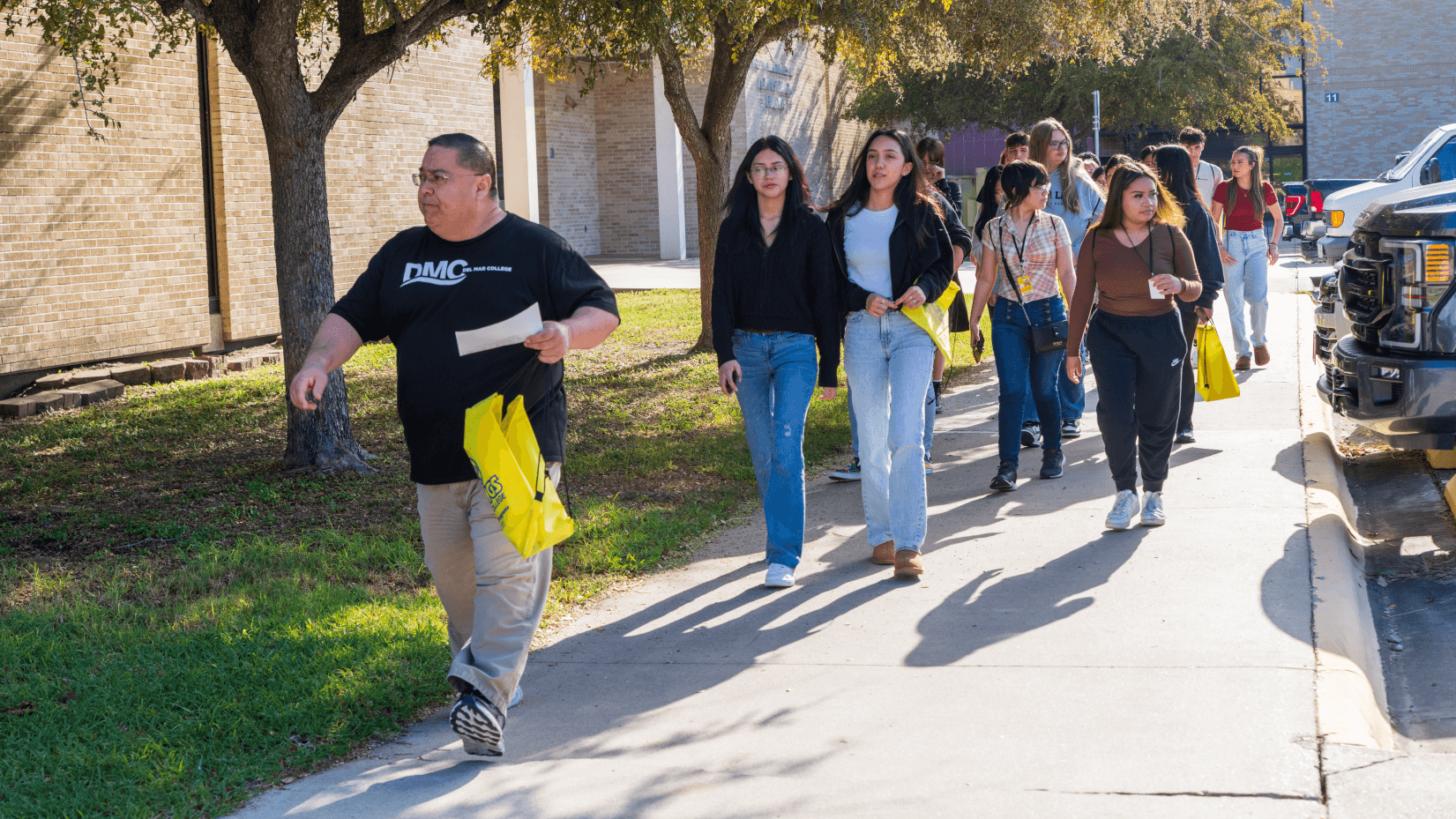 A group of people carrying yellow bags walks beside a brick building under tree shade.