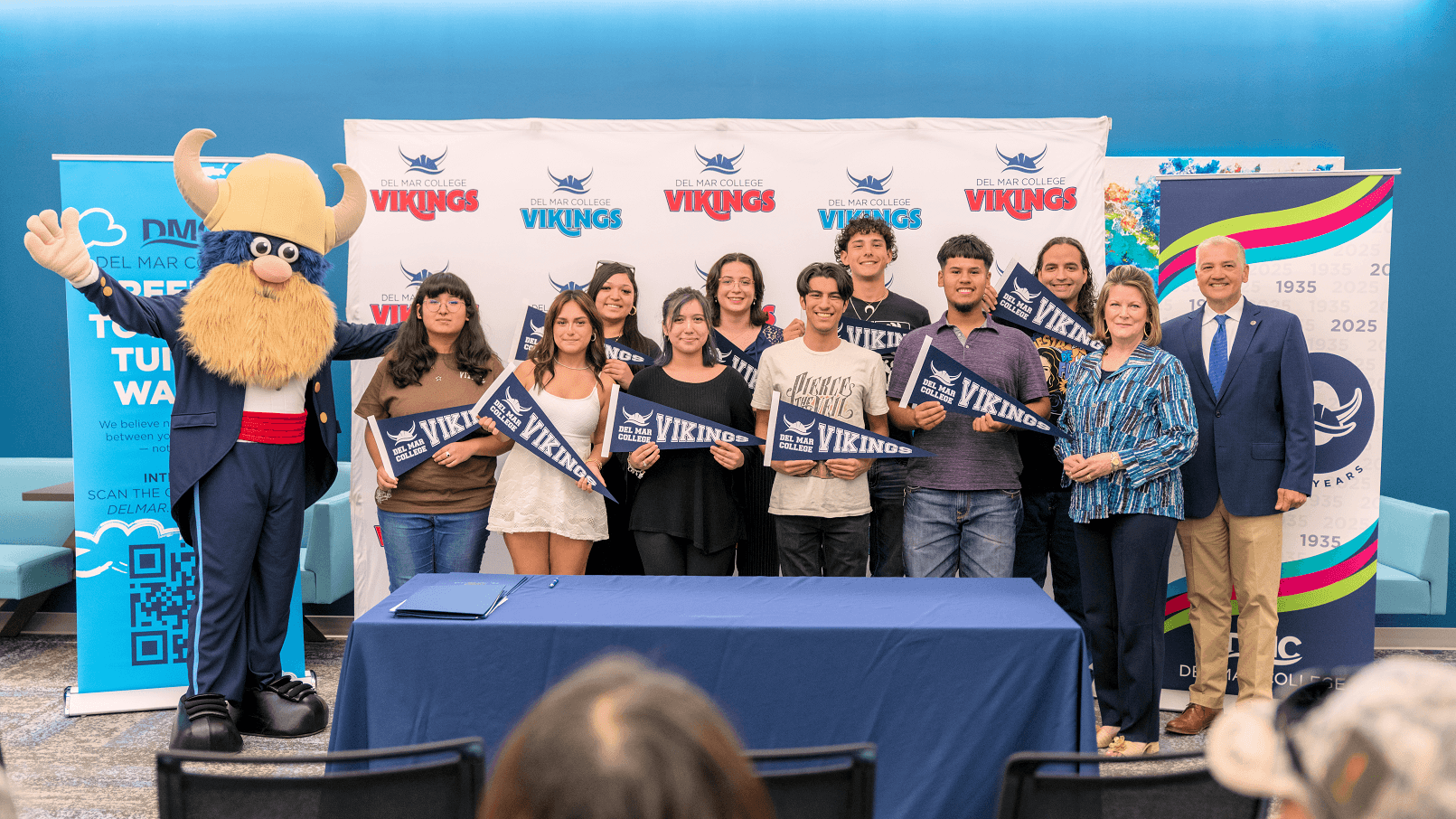 Group of students, college officials, and Valdar the Viking mascot pose with Del Mar College Vikings pennants at an indoor event.