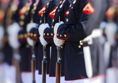 Line of uniformed individuals in dark blue military uniforms with red and gold accents, holding rifles.